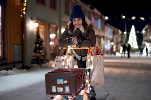 Eine Frau in Winterkleidung stützt sich nachts auf einer verschneiten Straße auf einen Schlitten, der mit Lichtern geschmückt und mit Weihnachtseinkaufstüten und einer braunen Schachtel gefüllt ist.