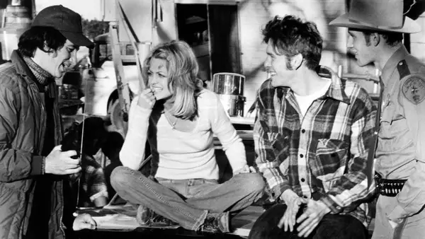 A black-and-white photo of a man wearing a jacket and a hat, three actors on the right with their attention to him as he speaks. She sits cross-legged on the hood of a truck, the man to her right smiling, the man on the far right dressed as law enforcement.