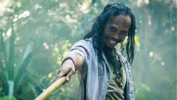 A man in the woods with with long, ragged hair smiles and holds out a fighting stick.