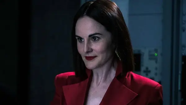 A woman dressed in a striking red formal suit smirks at something off camera.