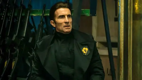 A man dresseded in an authoritarian-looking but stylish black suit with a yellow flower on the lapel stares at something out of frame