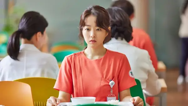 A young woman in scrubs sits at a cafeteria table with a distant and forlorn look on her face