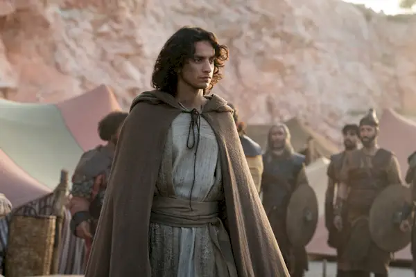 Ein Mann mit dunklen Locken und einem braunen Kapuzenumhang steht in einem Lager.